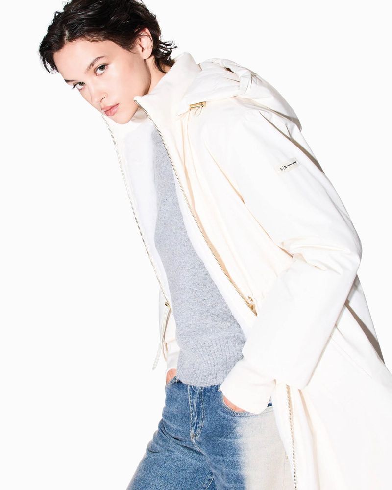 Fashion shot of a slender, fair-skinned woman in a long, white hooded coat and blue jeans against a stark white backdrop in a studio setting.