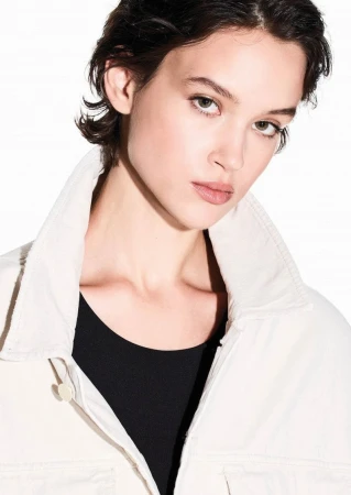 Close-up portrait of a woman with short dark hair wearing an off-white collared jacket over a black top against a white background.