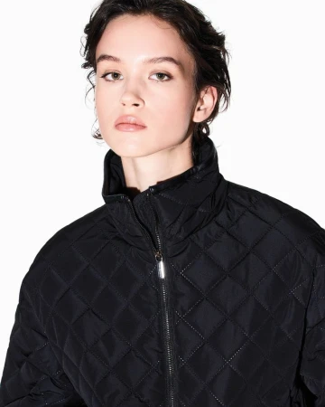 A woman with short dark hair wearing a black diamond-quilted zip jacket against a plain white studio background.