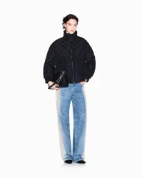 Model wearing a black quilted jacket and faded wide-leg denim jeans holding a black handbag against a white studio backdrop.