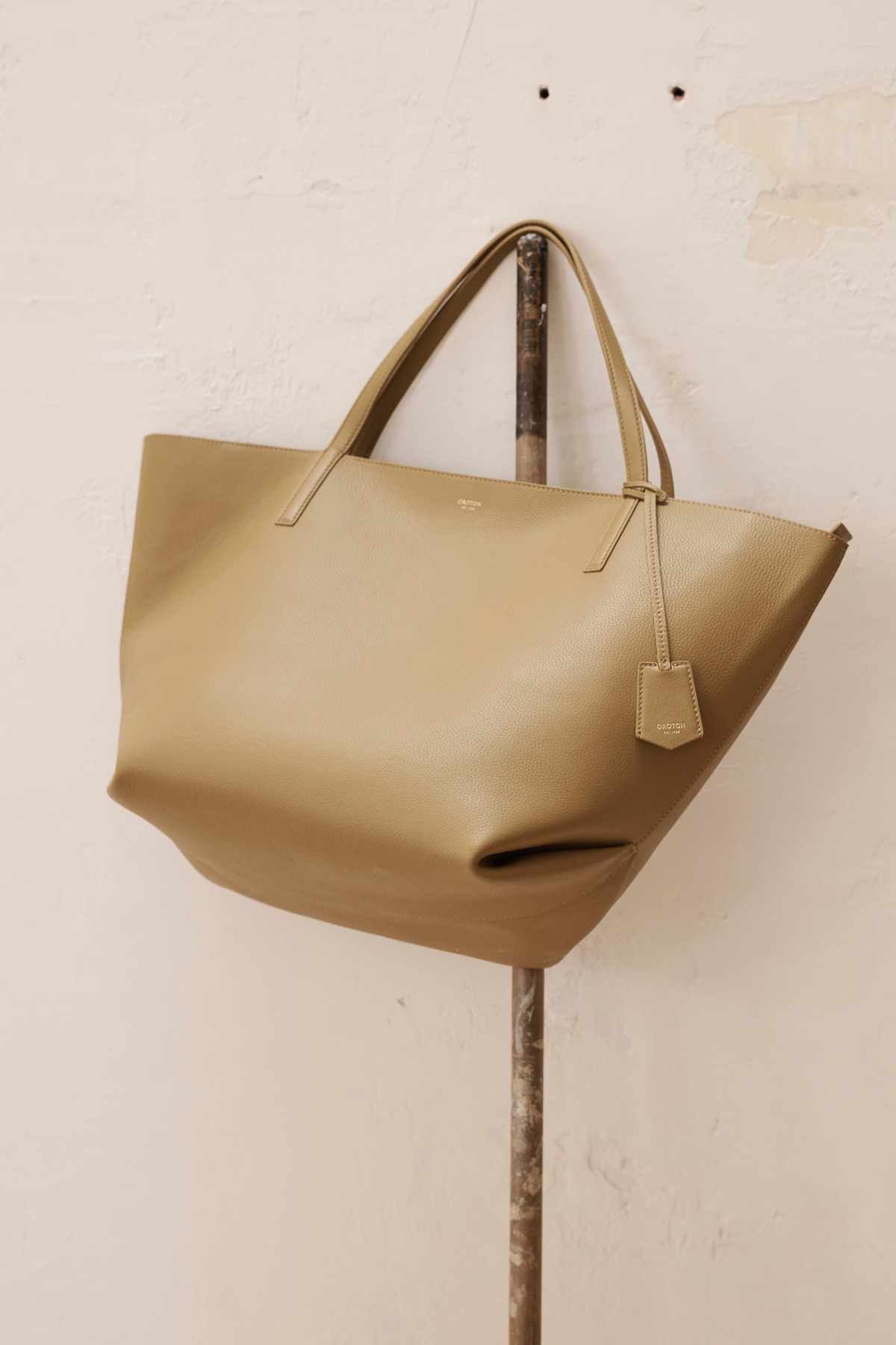 Beige leather tote bag hanging from a metal rod against a cream wall, studio setting, displaying fashion accessory.