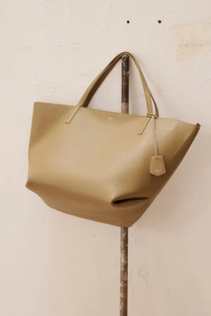 Beige leather tote bag hanging from a metal rod against a cream wall, studio setting, displaying fashion accessory.