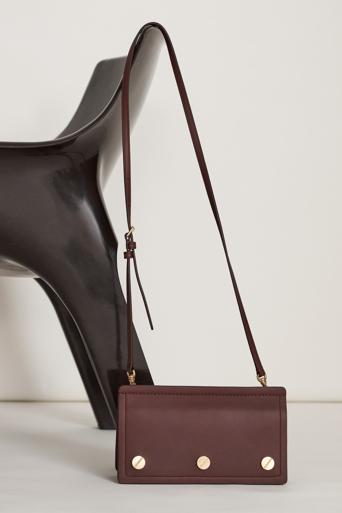 Burgundy-colored crossbody bag with gold hardware on a white surface, complemented by a dark brown modern chair in the background.