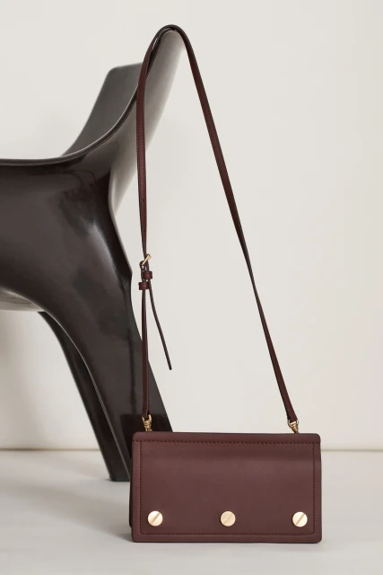 Burgundy-colored crossbody bag with gold hardware on a white surface, complemented by a dark brown modern chair in the background.