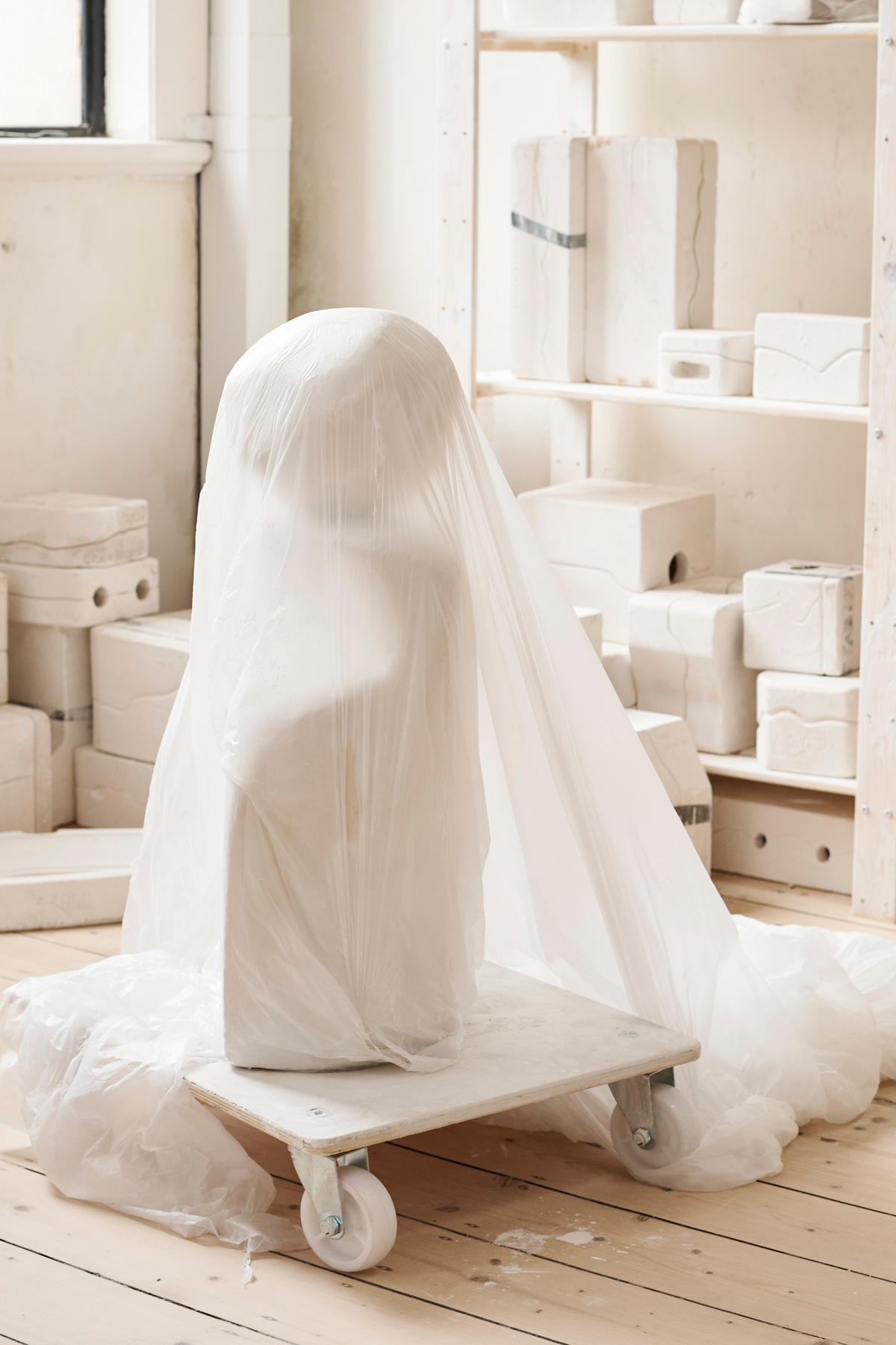 White sculpture covered in translucent plastic wrap on a wheeled platform in an artist's studio with white walls and display shelves.