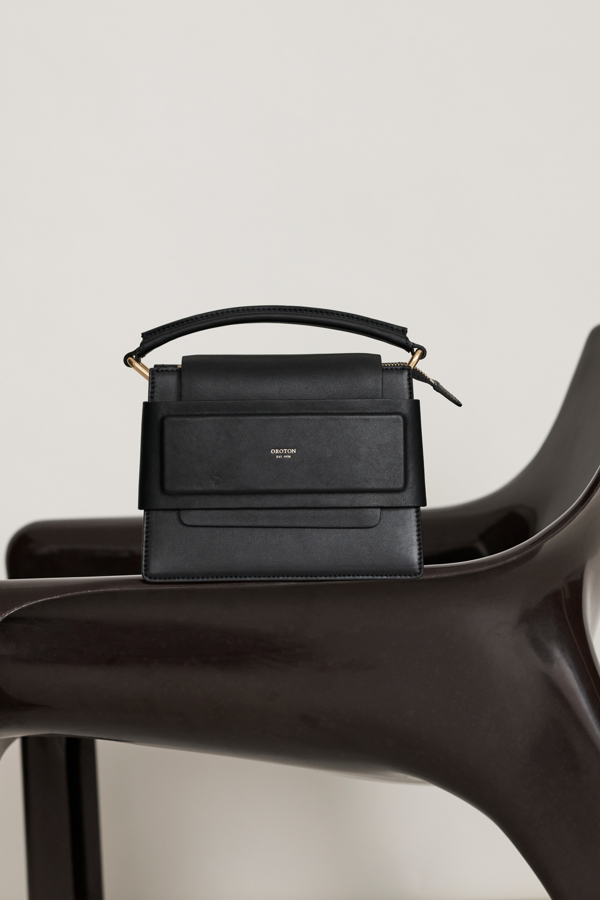 Black leather handbag with a handle on a modern wooden chair against a white background in a fashion product studio shot.