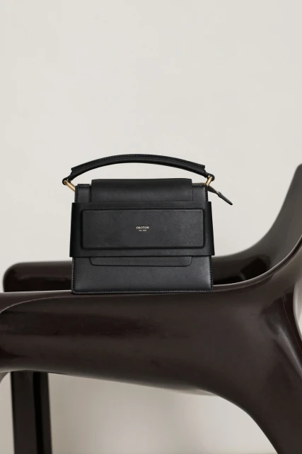 Black leather handbag with a handle on a modern wooden chair against a white background in a fashion product studio shot.