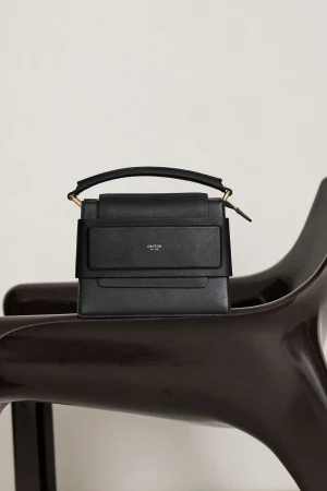 Black leather handbag with a handle on a modern wooden chair against a white background in a fashion product studio shot.