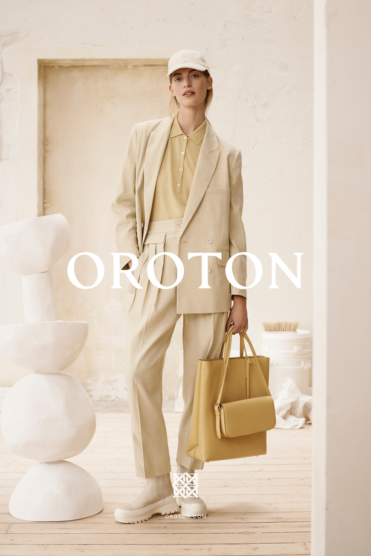Fashion model wearing a light beige blazer and trouser suit, accessorized with a cap and handbag, poses in an indoor setting.