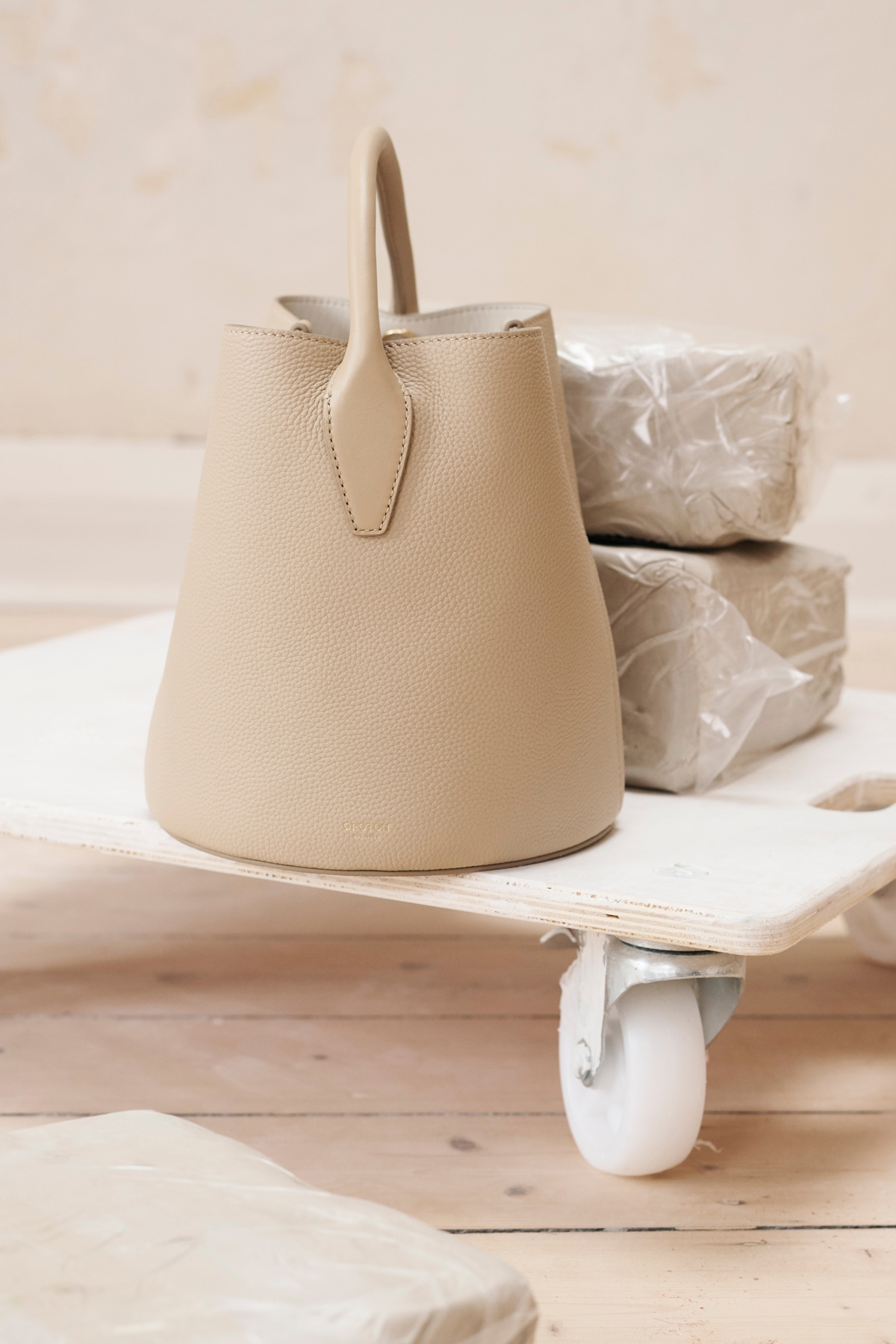 Beige bucket bag with handle on a white wheeled platform, alongside wrapped clay blocks, against a light beige background in a studio.