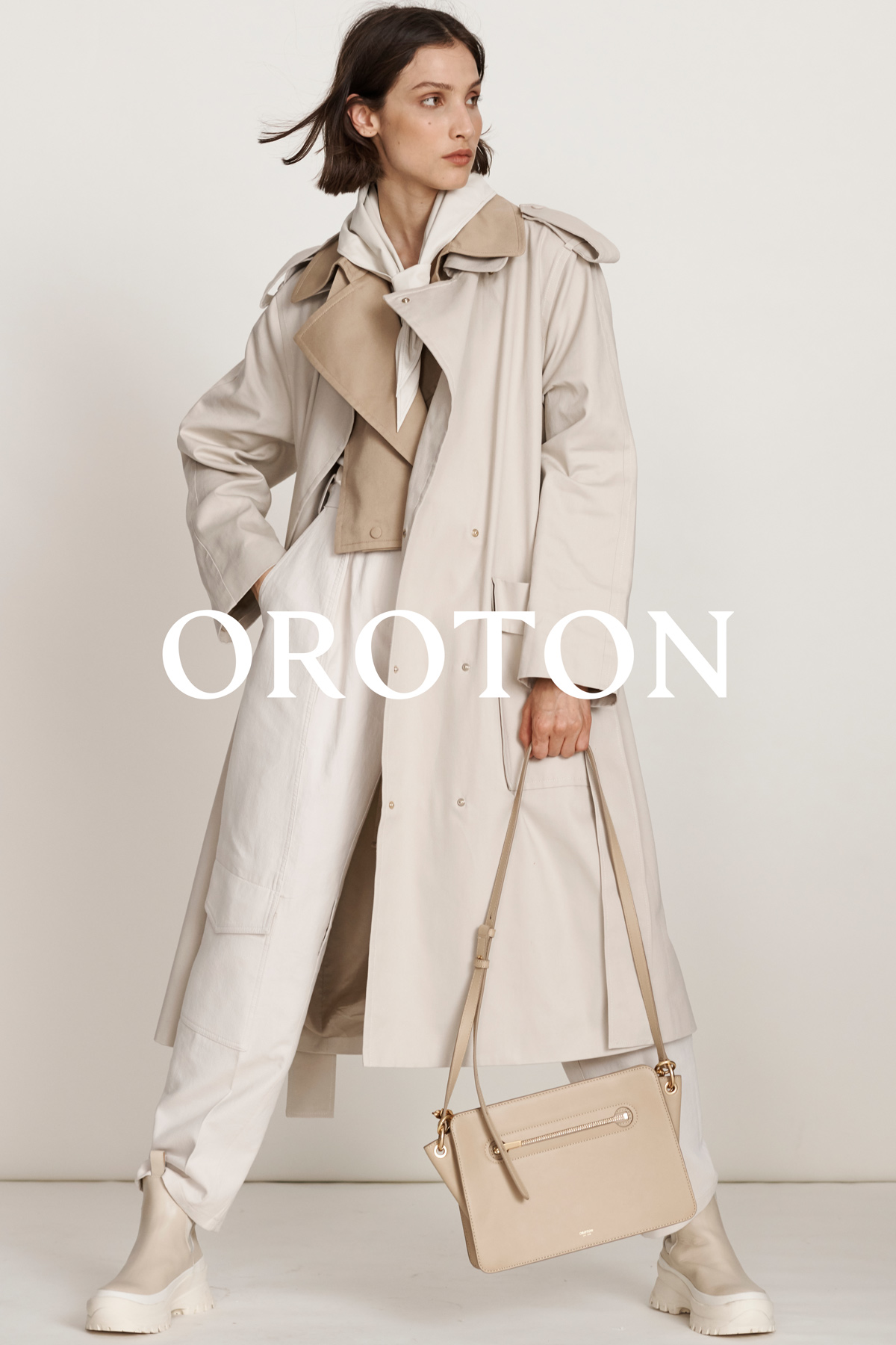 Fashion shot of a woman in a tan trench coat holding a beige handbag, standing against a plain background in a studio setting.