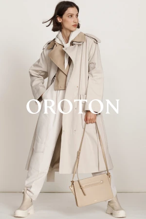 Fashion shot of a woman in a tan trench coat holding a beige handbag, standing against a plain background in a studio setting.