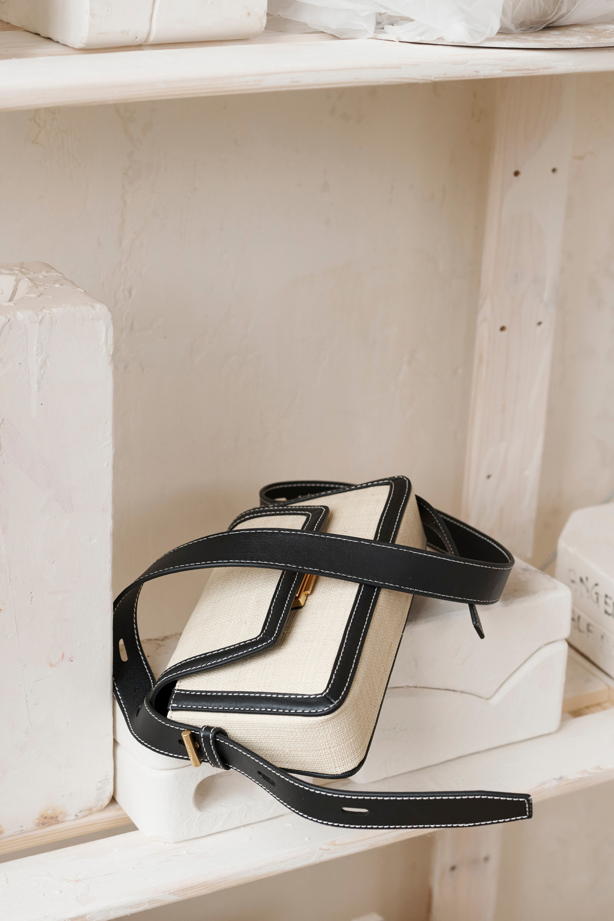 Still life shot of a cream-colored purse with black straps and trim, resting on a white shelf with other plaster objects nearby.