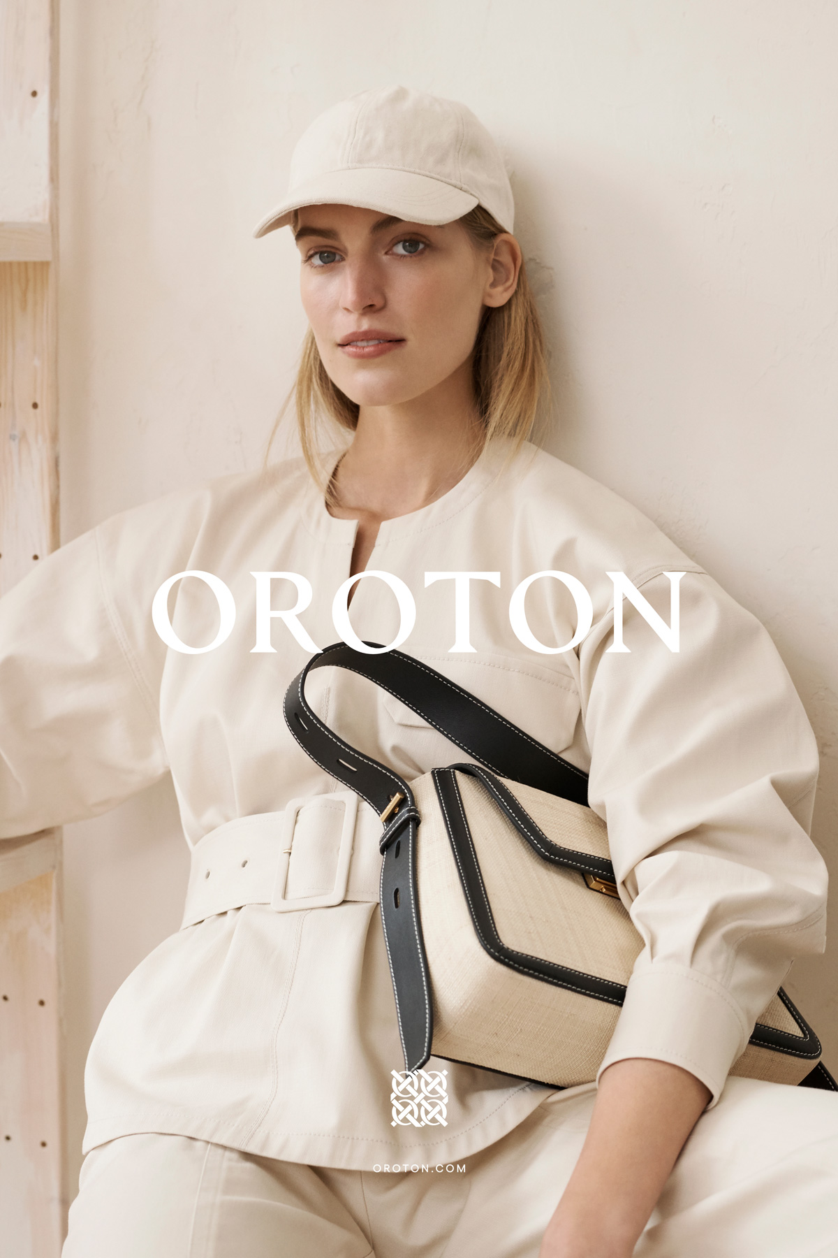 Model Vanessa Axente wearing cream jacket and cap holding Oroton bag against a neutral wall