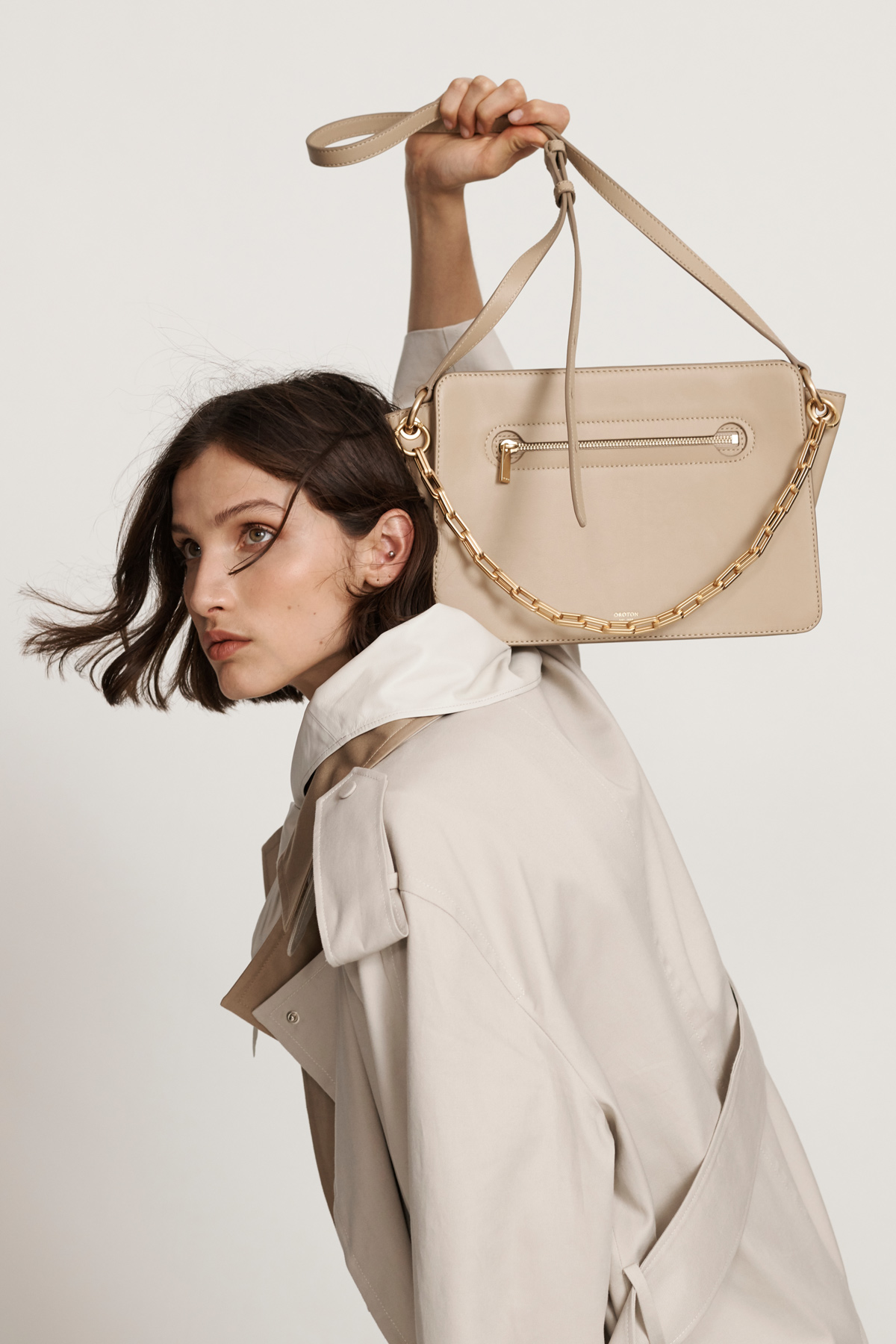 Woman showcases a beige handbag with a chain strap in a studio setting, wearing a light trench coat against a neutral background.