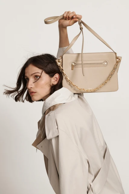 Woman showcases a beige handbag with a chain strap in a studio setting, wearing a light trench coat against a neutral background.