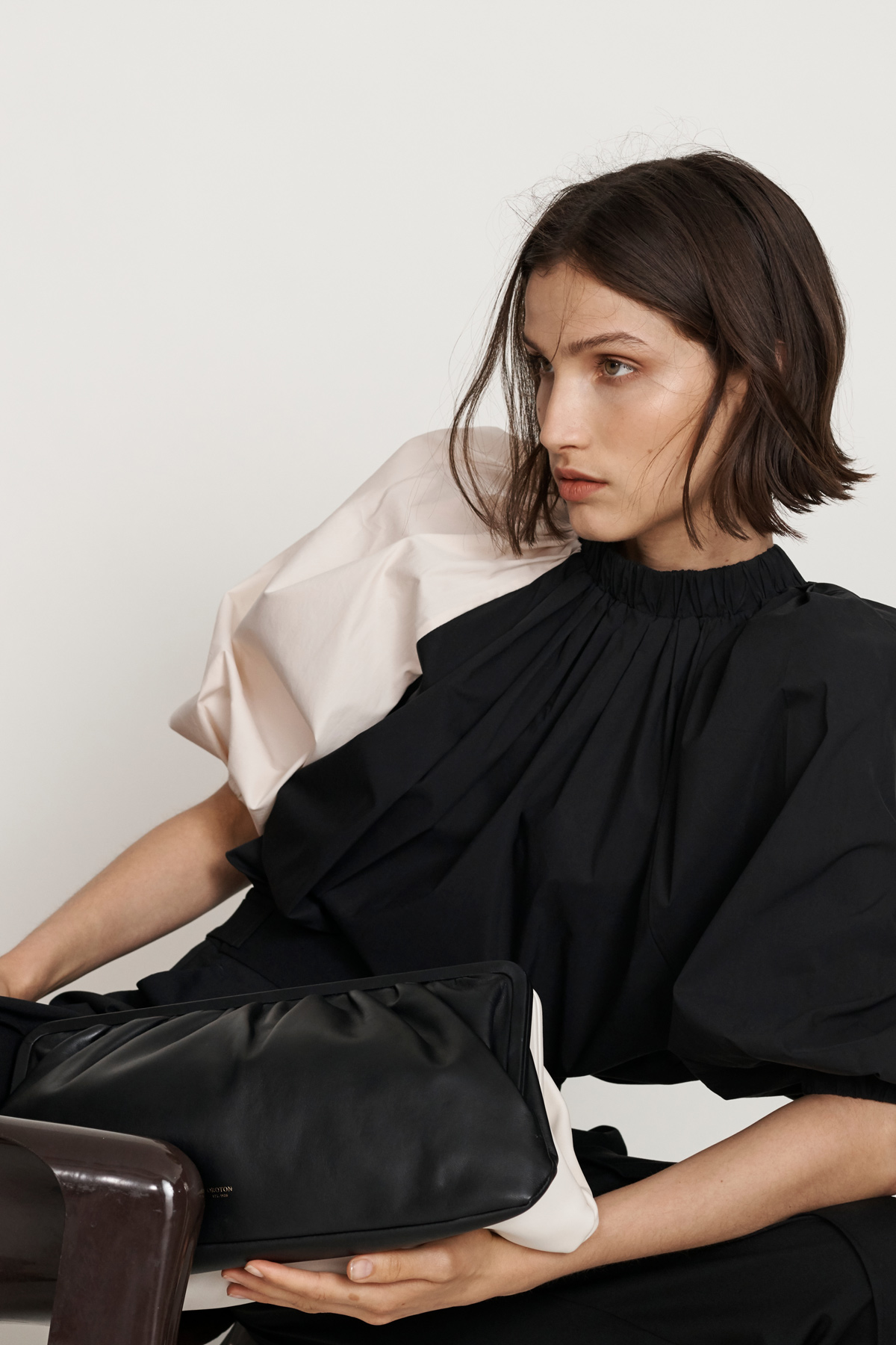 A brunette woman wearing a black top with a pale pink puffy sleeve poses with a black leather clutch bag against a white background.