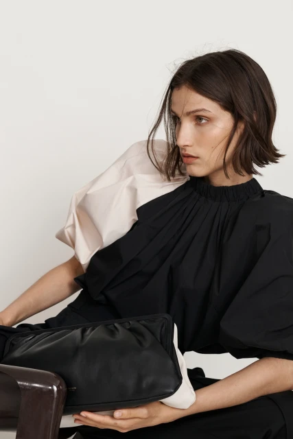 A brunette woman wearing a black top with a pale pink puffy sleeve poses with a black leather clutch bag against a white background.