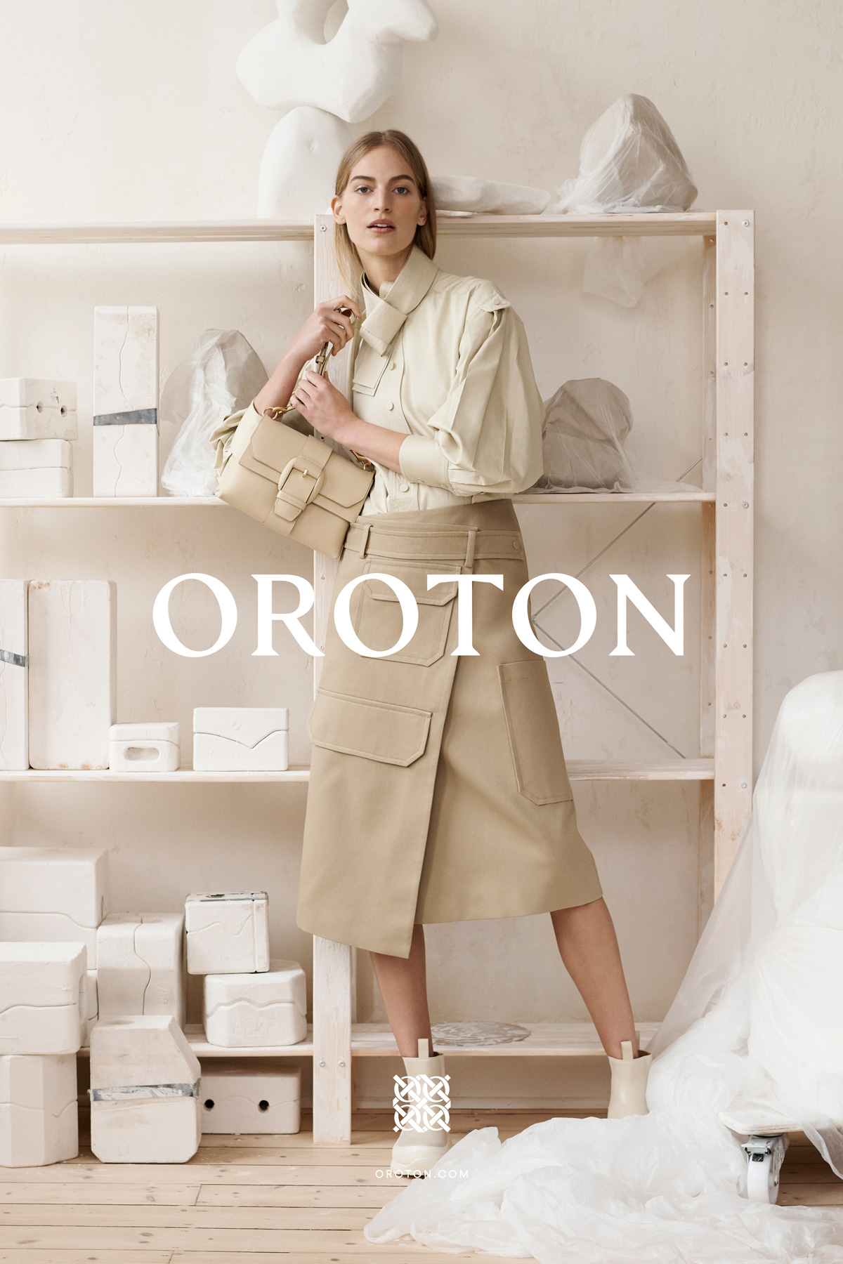 Fashion editorial shot of a woman in a tan skirt and shirt holding a purse in a light-filled studio with abstract art.
