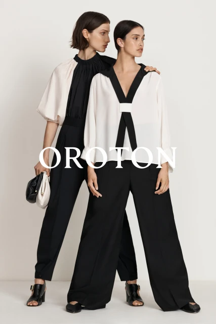Two fashion models pose in black and white blouses with wide-legged black pants against a minimalist background, showcasing modern elegance.