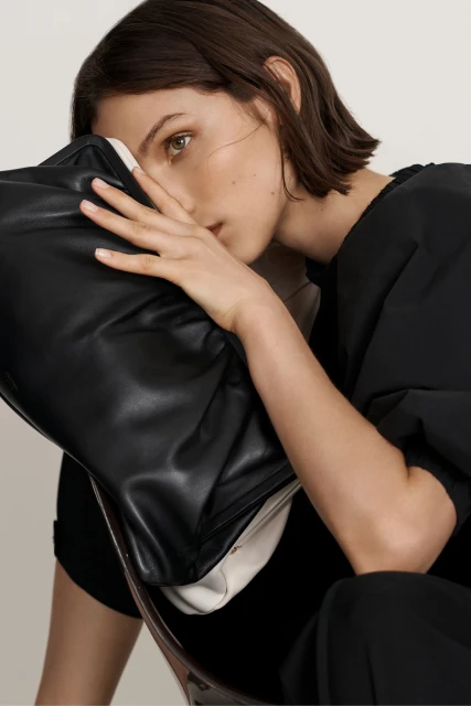 Adult woman in dark clothing holding a black leather bag, partially obscuring her face, indoors, with neutral light.
