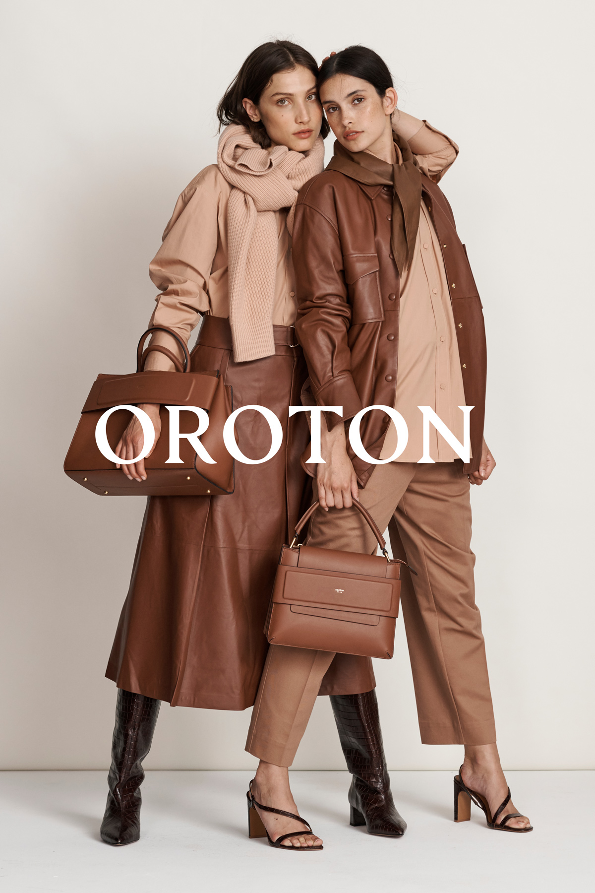 Two women pose indoors modeling Oroton leather handbags, wearing tan-colored outfits with high-heeled sandals and knee-high boots.