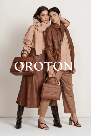 Two women pose indoors modeling Oroton leather handbags, wearing tan-colored outfits with high-heeled sandals and knee-high boots.