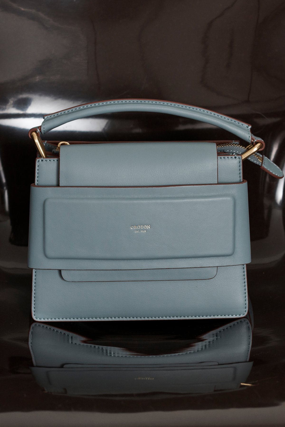Studio shot of a light blue leather handbag with a top handle and gold hardware, reflecting on a dark, shiny surface.