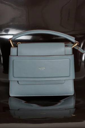 Studio shot of a light blue leather handbag with a top handle and gold hardware, reflecting on a dark, shiny surface.