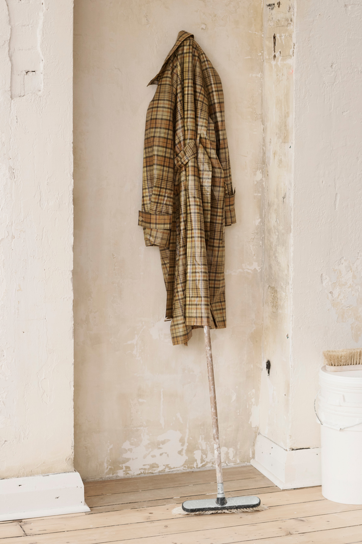 Plaid trench coat hangs on a wall above a floor cleaning mop in a room with white painted walls and wooden flooring.