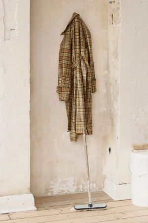 Plaid trench coat hangs on a wall above a floor cleaning mop in a room with white painted walls and wooden flooring.