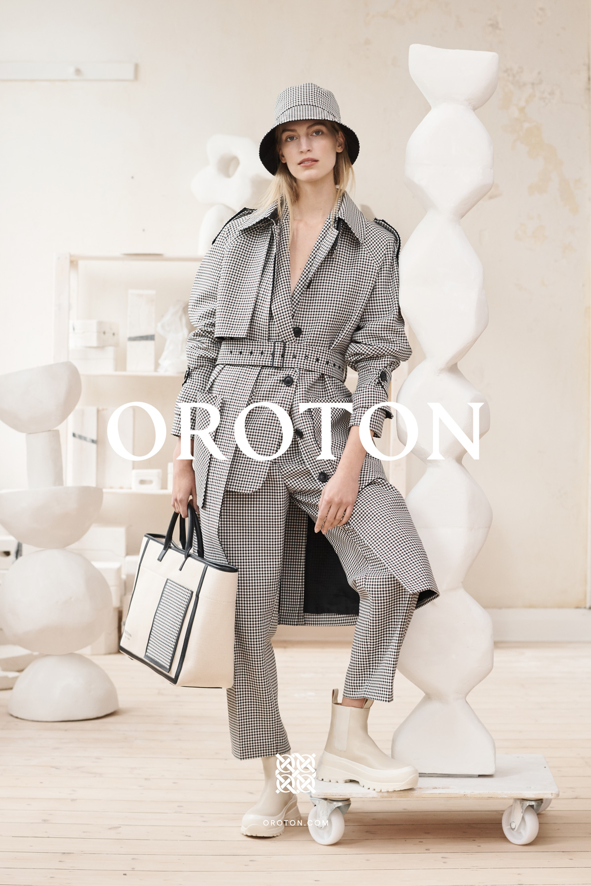 A woman in a plaid trench coat and bucket hat, holding a handbag, stands on a platform in a fashion editorial shot indoors.