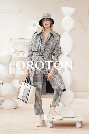 A woman in a plaid trench coat and bucket hat, holding a handbag, stands on a platform in a fashion editorial shot indoors.