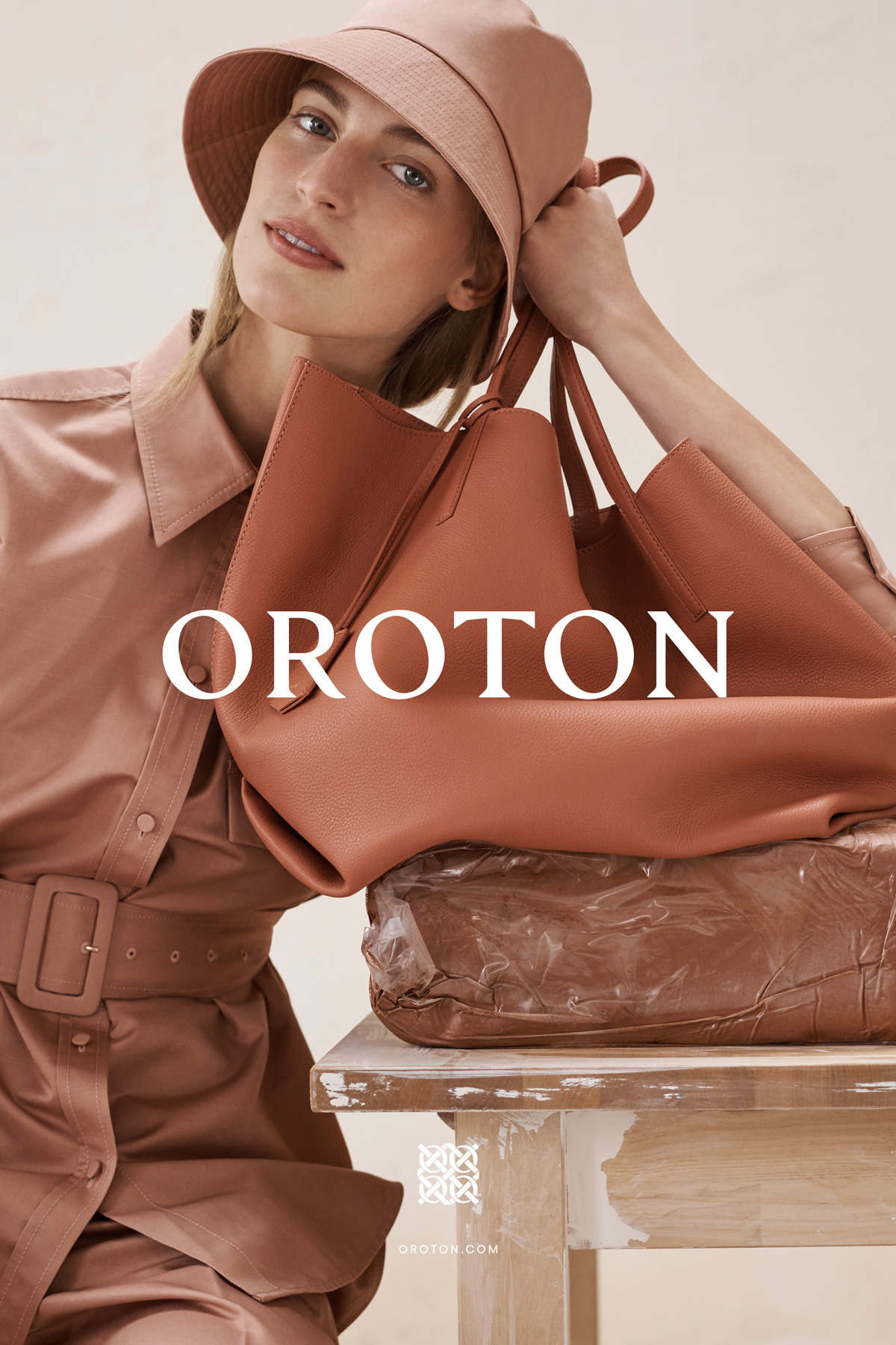 Blonde woman in salmon-colored outfit poses indoors with a large salmon-colored leather handbag, sitting on a white painted table.