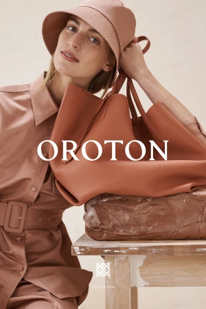 Blonde woman in salmon-colored outfit poses indoors with a large salmon-colored leather handbag, sitting on a white painted table.