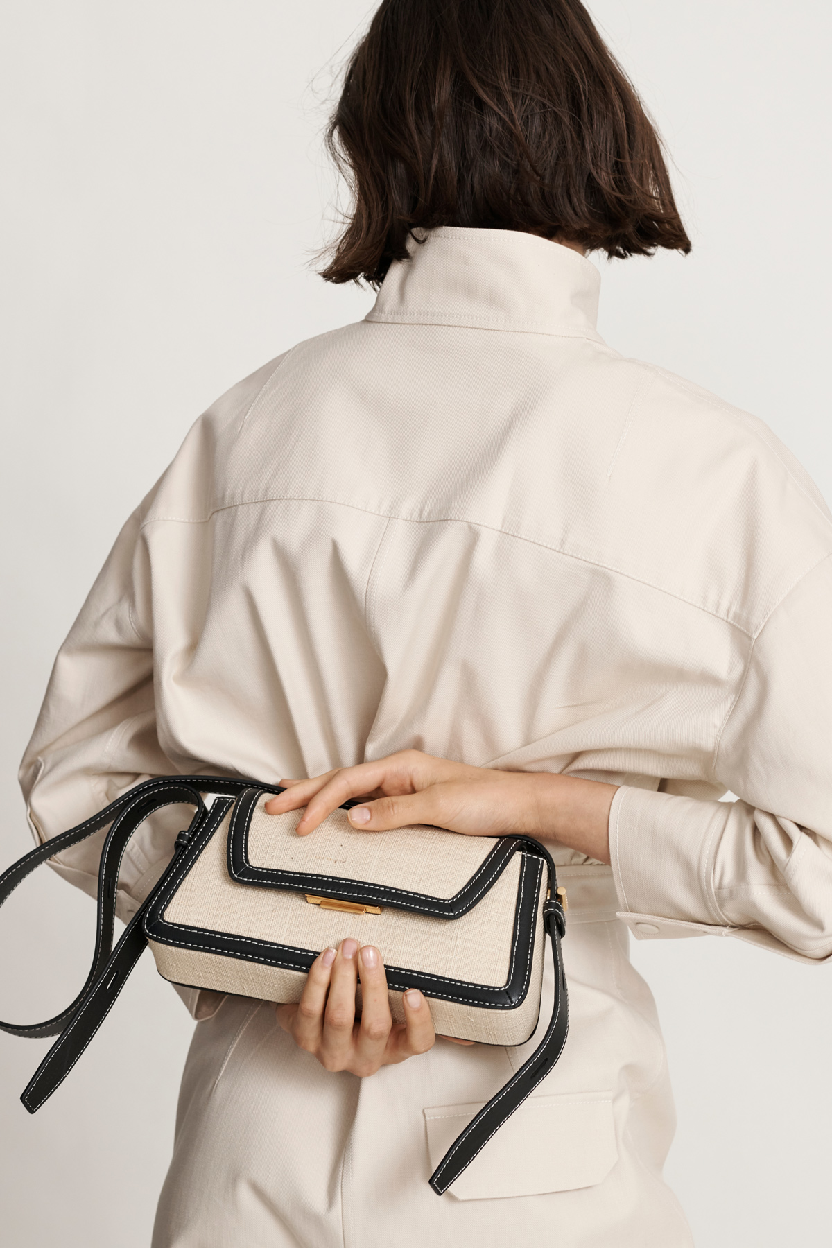 Woman showcases a cream-colored cross-body bag with black trim and strap against a white backdrop, complementing her matching cream jumpsuit.
