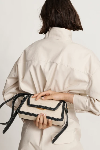 Woman showcases a cream-colored cross-body bag with black trim and strap against a white backdrop, complementing her matching cream jumpsuit.