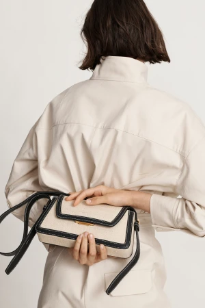 Woman showcases a cream-colored cross-body bag with black trim and strap against a white backdrop, complementing her matching cream jumpsuit.