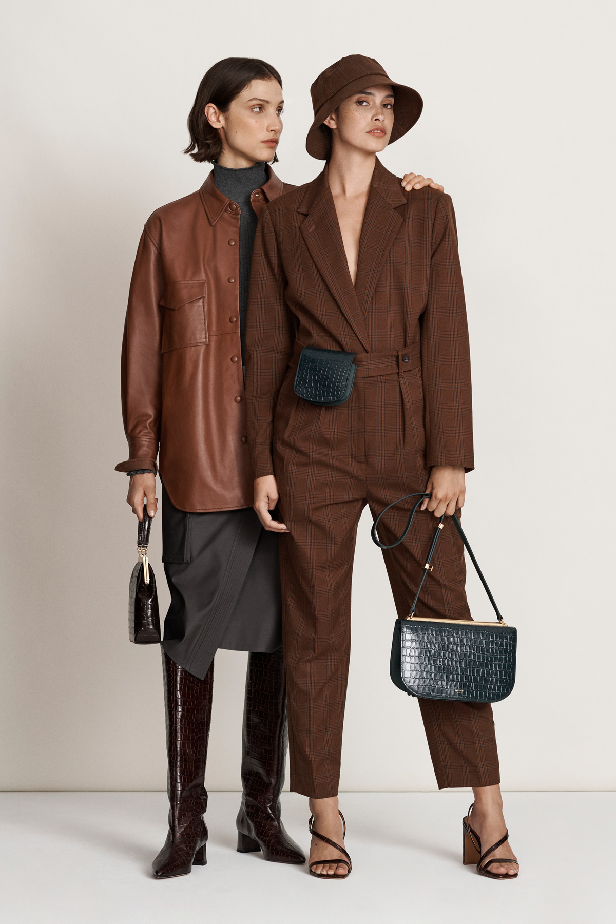 Two women pose in brown and grey outfits in a studio setting. One woman wears a brown jacket and boots, the other a brown suit and hat.