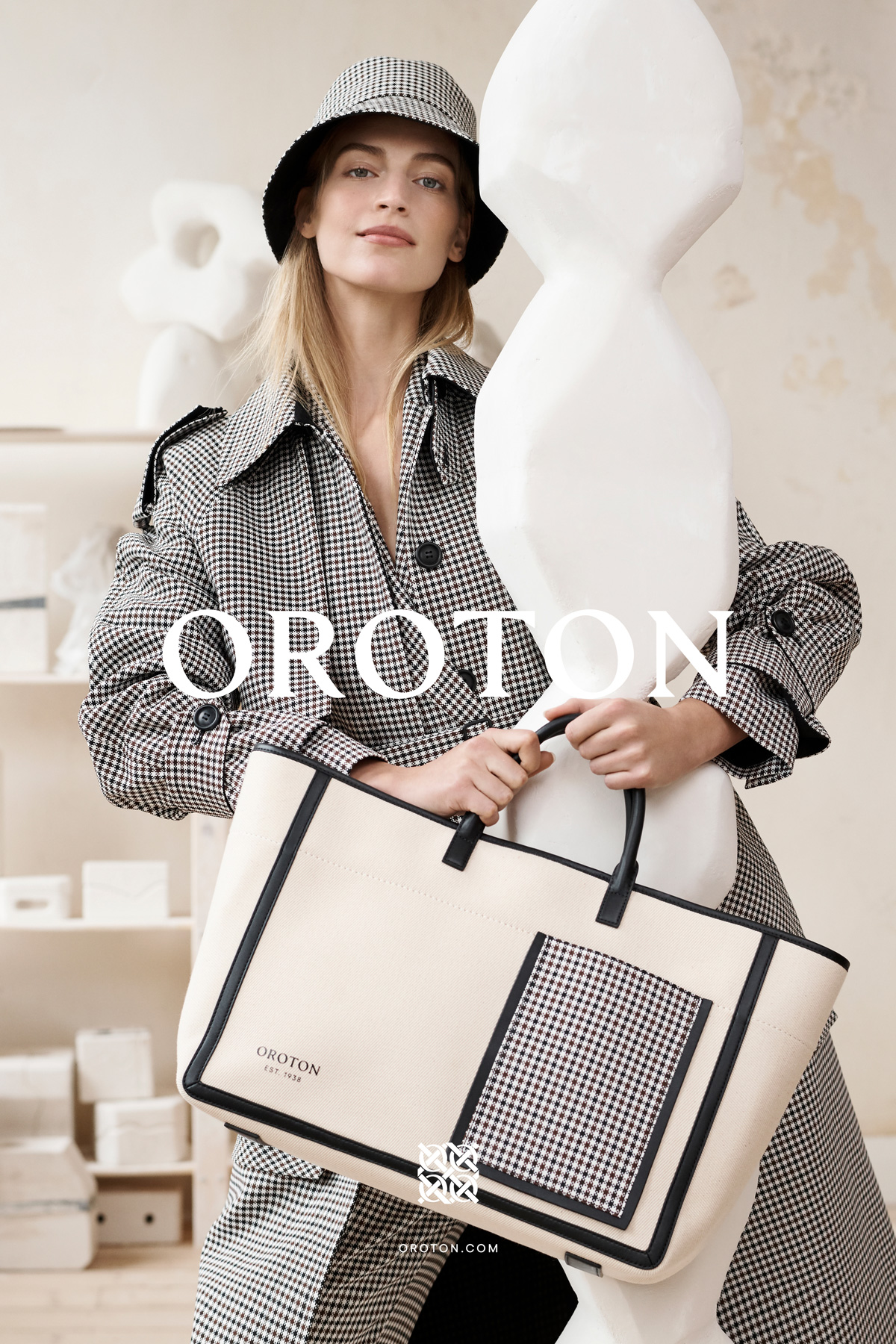 A blonde adult female model poses indoors holding a large tote bag, wearing a black and white houndstooth coat and matching hat.