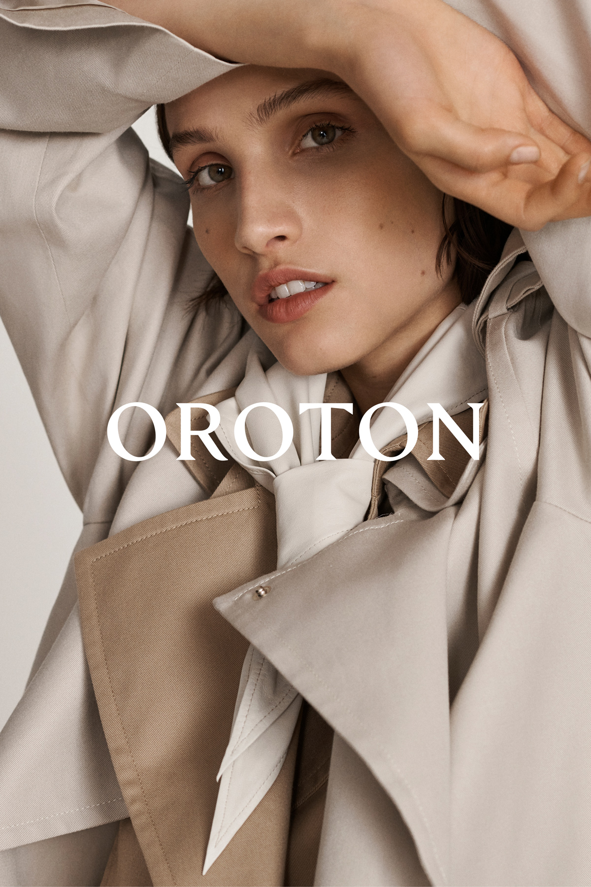 Close-up of a woman with a neutral expression in a light beige trench coat, with the brand name OROTON overlayed on the image.