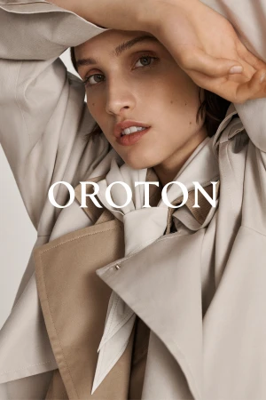 Close-up of a woman with a neutral expression in a light beige trench coat, with the brand name OROTON overlayed on the image.