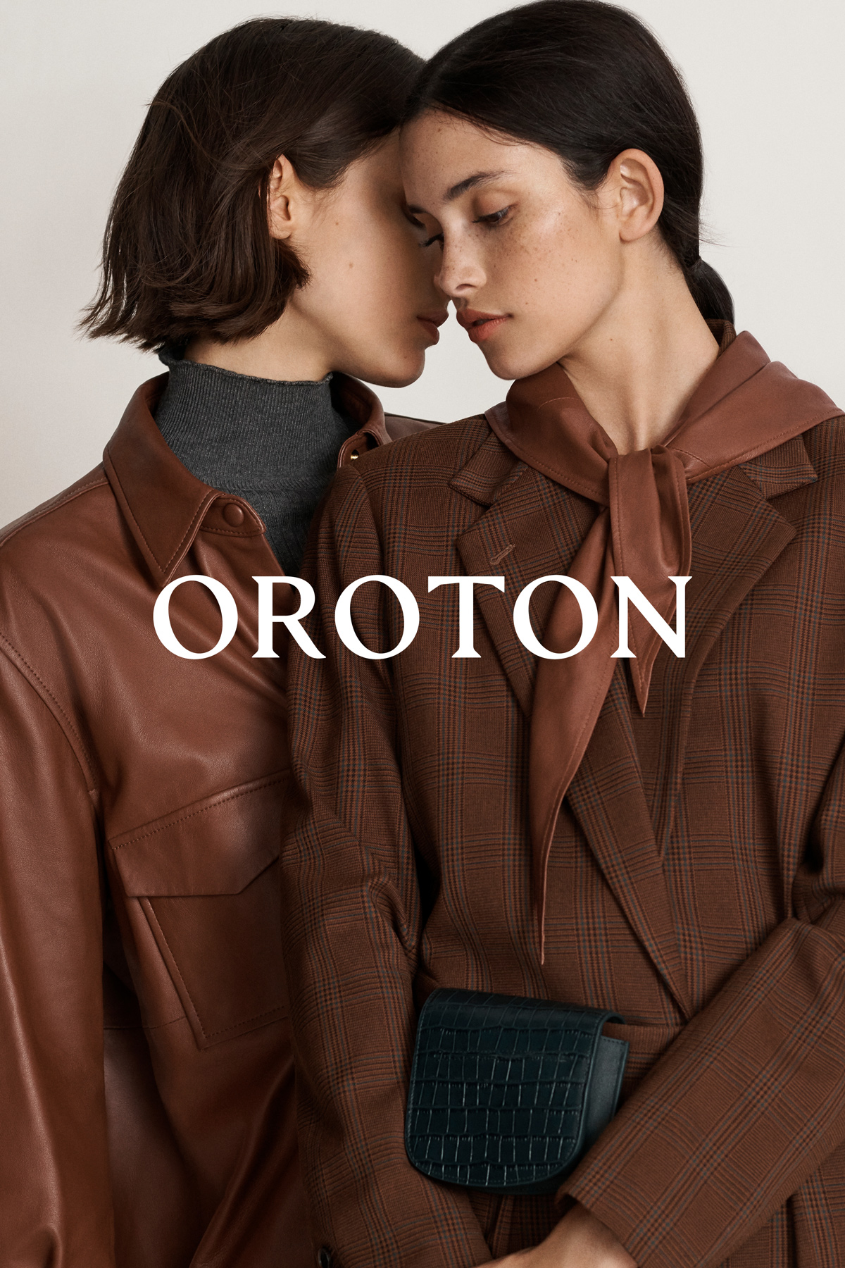 Two adult women pose in a fashion shot, showcasing brown jackets in an indoor setting with the brand name Oroton displayed.
