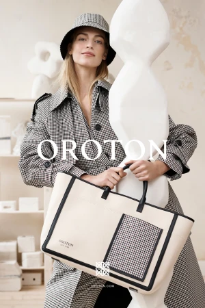 Model in houndstooth coat and matching bucket hat holding a cream tote bag with black trim in a white studio setting.