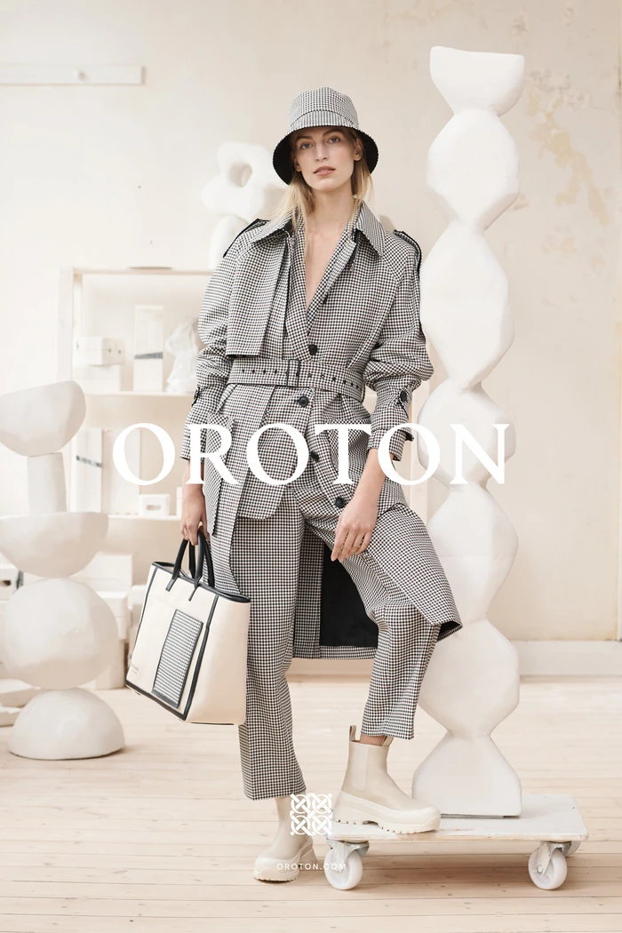 Model in houndstooth trench coat and bucket hat holding a structured tote bag in an art-filled studio setting.