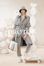 Model in houndstooth trench coat and bucket hat holding a structured tote bag in an art-filled studio setting.