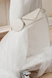 White leather belt bag with round and envelope pouches worn against crumpled white plastic and a wooden floor