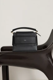 Black leather handbag resting on a dark brown molded chair against a plain light wall