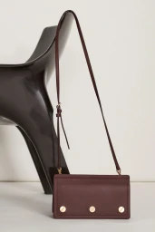 Burgundy leather crossbody bag with metal rivets positioned in front of a dark glossy sculptural chair against a white background.