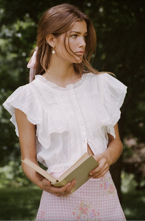 Woman in white blouse with ruffles holds a book outdoors, with a pink ribbon in her hair and a blurred green background.